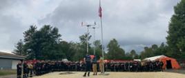 Poranna odprawa grupy na tle obozowiska. Centalnie widoczny masz z polską flagą oraz maszty samochodu dowodzenia i łączności