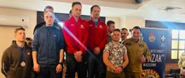 Kolorowa fotografia wykonana w pomieszczeniu po zawodach pływackich strażaków PSP. Na pierwszym planie medaliści stojący na podium, obok nich znajdują się strażacy w mundurach, w tle banery Komendy Wojewódzkiej PSP i klubu pożarniczego Strażak.
