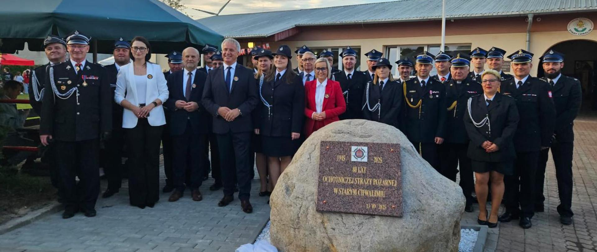 Na zdjęciu widać dużą grupę osób, w większości strażaków w galowych mundurach, zebranych wokół dużego kamienia pamiątkowego. Kamień ma przymocowaną brązową tablicę z napisem: "80 LAT OCHOTNICZEJ STRAŻY POŻARNEJ W STARYM CHWALIMIE". W pierwszym rzędzie, pośrodku, stoi kilku strażaków oraz dwie kobiety w białych bluzkach i ciemnych marynarkach. W tle znajduje się budynek straży pożarnej, na dachu którego widać fragment turbiny wiatrowej. Nad budynkiem wisi polska flaga. Niebo jest lekko zachmurzone, oświetlone promieniami zachodzącego słońca.