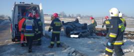 Na zdjęciu grupa strażaków stojących obok uszkodzonego pojazdu. Obok widać karetkę pogotowia.