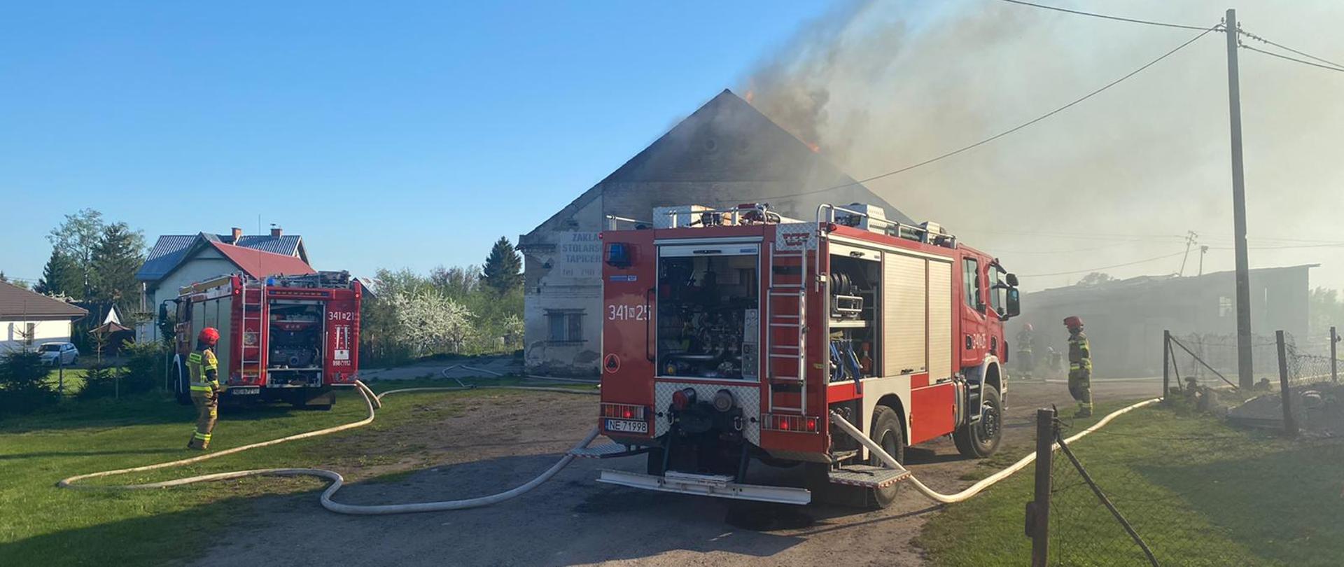 Na pierwszym planie dwa pojazdy pożarnicze, w tle widoczny budynek gospodarczy, którego dach w całości jest objęty pożarem, widoczny jest ogień i dym.