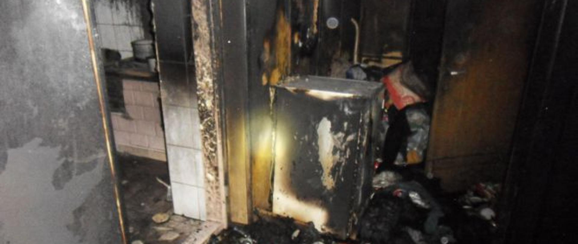 Zdjęcie przedstawia spalone pomieszczenie budynku mieszkalnego. W tle widoczny sprzęt AGD, pomieszczenie kuchenne oraz duża ilość materiału palnego zgromadzona w pomieszczeniu.