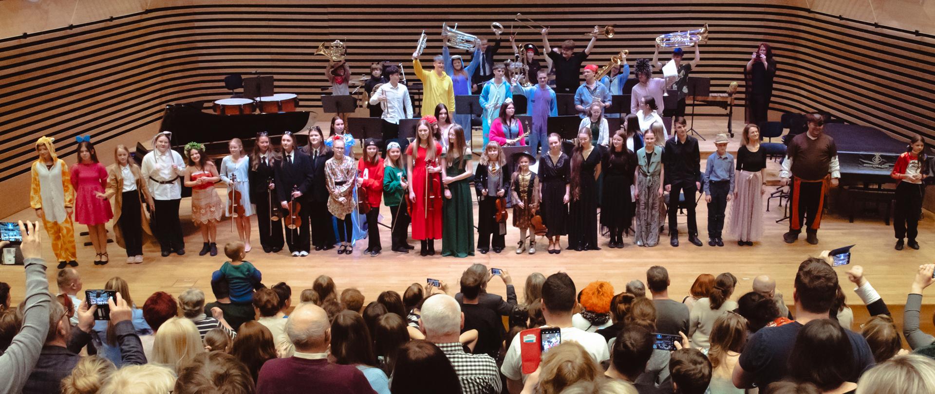 Wszyscy wykonawcy stoją na scenie, z przodu widać od tyłu stojącą publiczność.