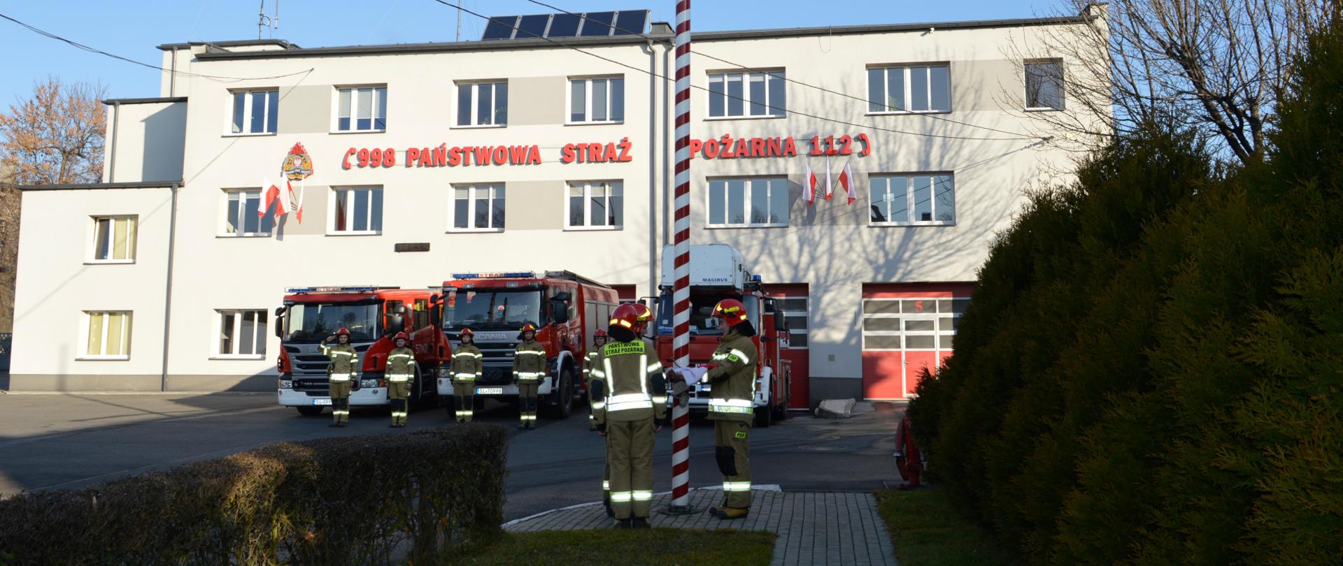 Na zdjęciu znajdują się strażacy w szeregu oraz poczet flagowy mocujący flagę państwową na maszt na tle samochodów pożarniczych