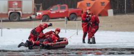 Strażacy wyciągają z przerębla poszkodowanego na ponton i ciągną linką na brzeg wraz ze strażakami w kombinezonach wypornościowych, w tle samochody strażackie