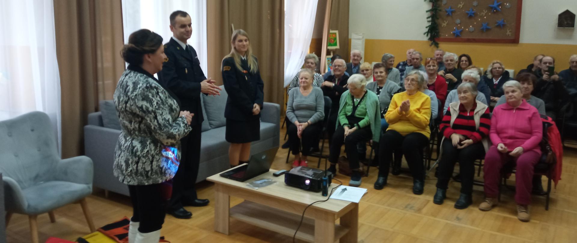 Sala wypełniona seniorami a przed nimi przemawia burmistrz Ćmielowa obok stoją funkcjonariusze PSP