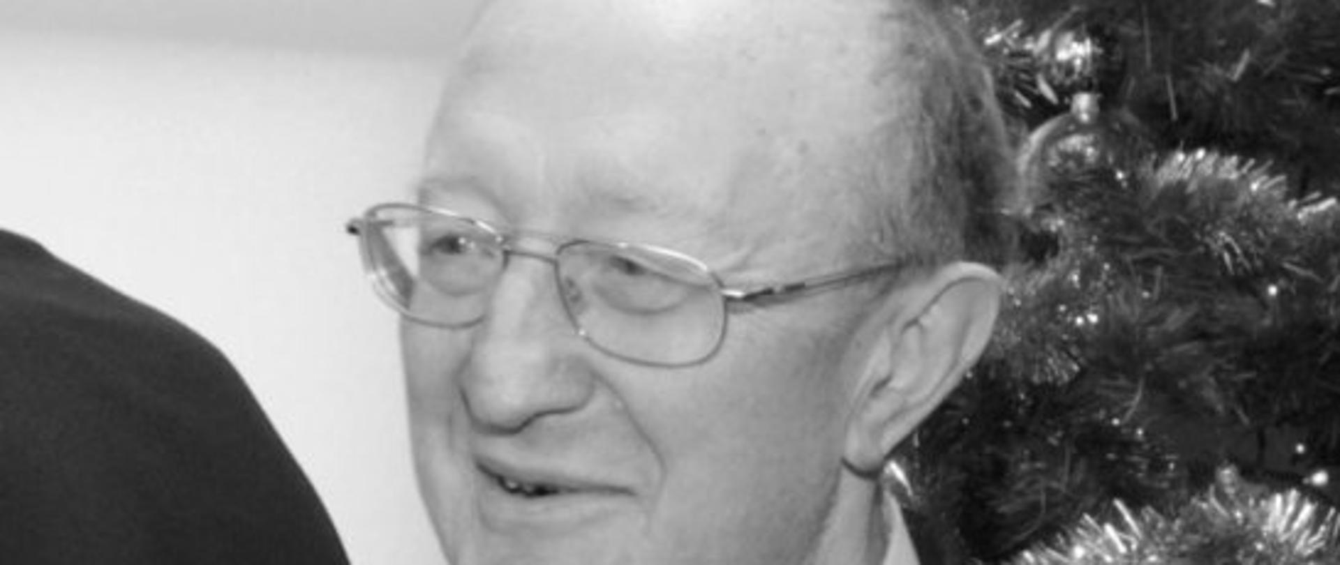 zdjęcie czarno białe przedstawia mężczyznę w okularach w garniturze w białej koszuli i z krawatem, w tle widać choinkę