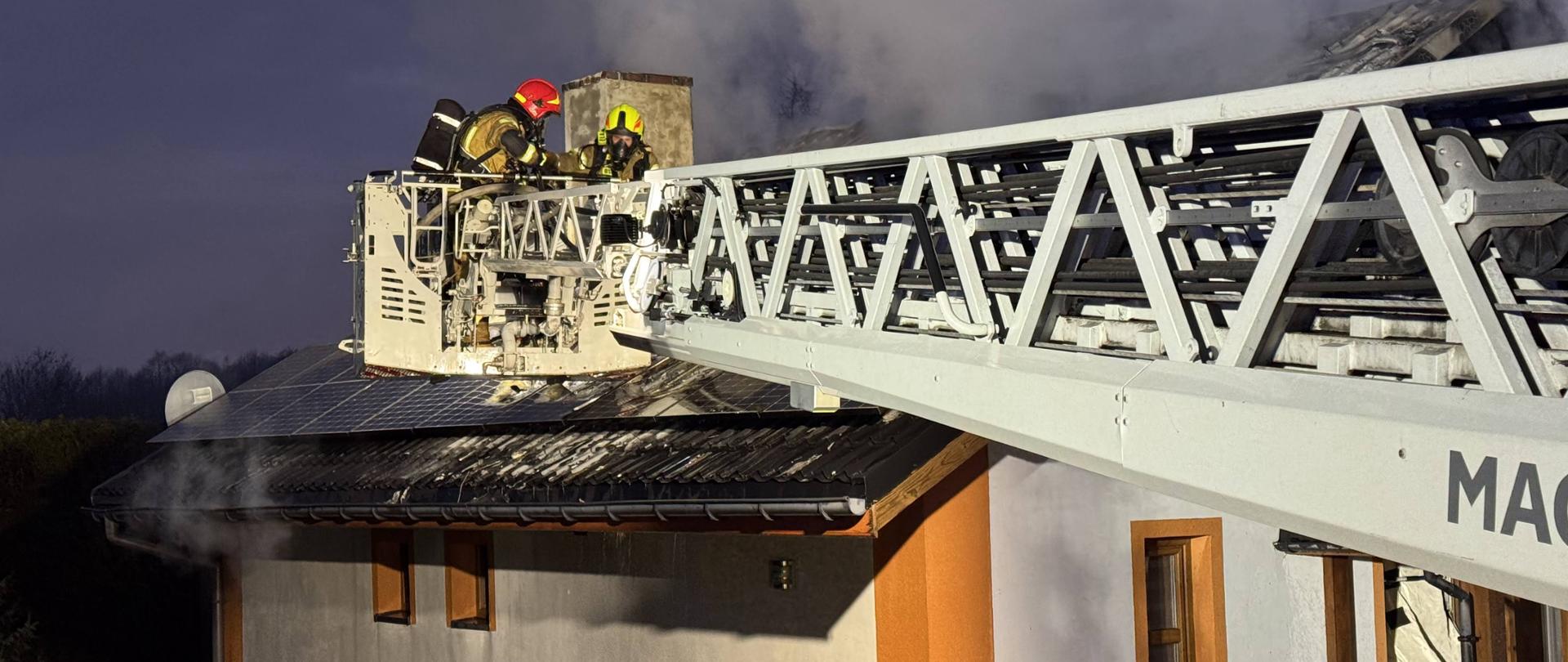 Strażacy w kaskach i pełnym umundurowaniu prowadzą akcję gaśniczą na dachu domu jednorodzinnego. Stoją na wysięgniku drabiny pożarniczej, zbliżając się do komina, z którego wydobywa się gęsty dym. Dach budynku jest częściowo uszkodzony, a na jego powierzchni widoczne są panele słoneczne. Scena ma miejsce o zmierzchu lub w nocy, a miejsce akcji oświetlone jest sztucznym światłem.