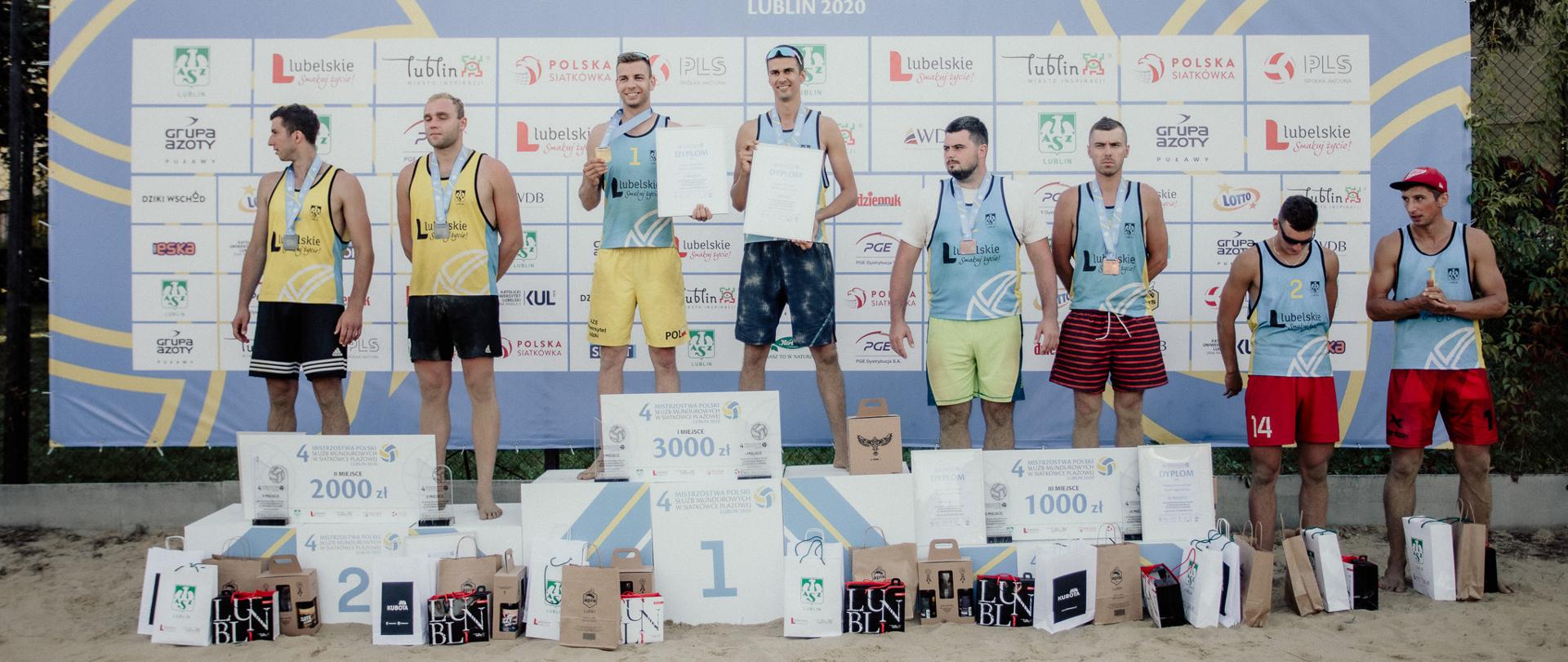 IV Mistrzostwa Polski Służb Mundurowych w Siatkówce Plażowej LUBLIN 2020 – na zdjęciu ośmiu zawodników na podium. Przed nimi nagrody rzeczowe oraz czeki na 1000, 2000 i 3000 zł. W tle tablica z reklamami firm sponsorów.