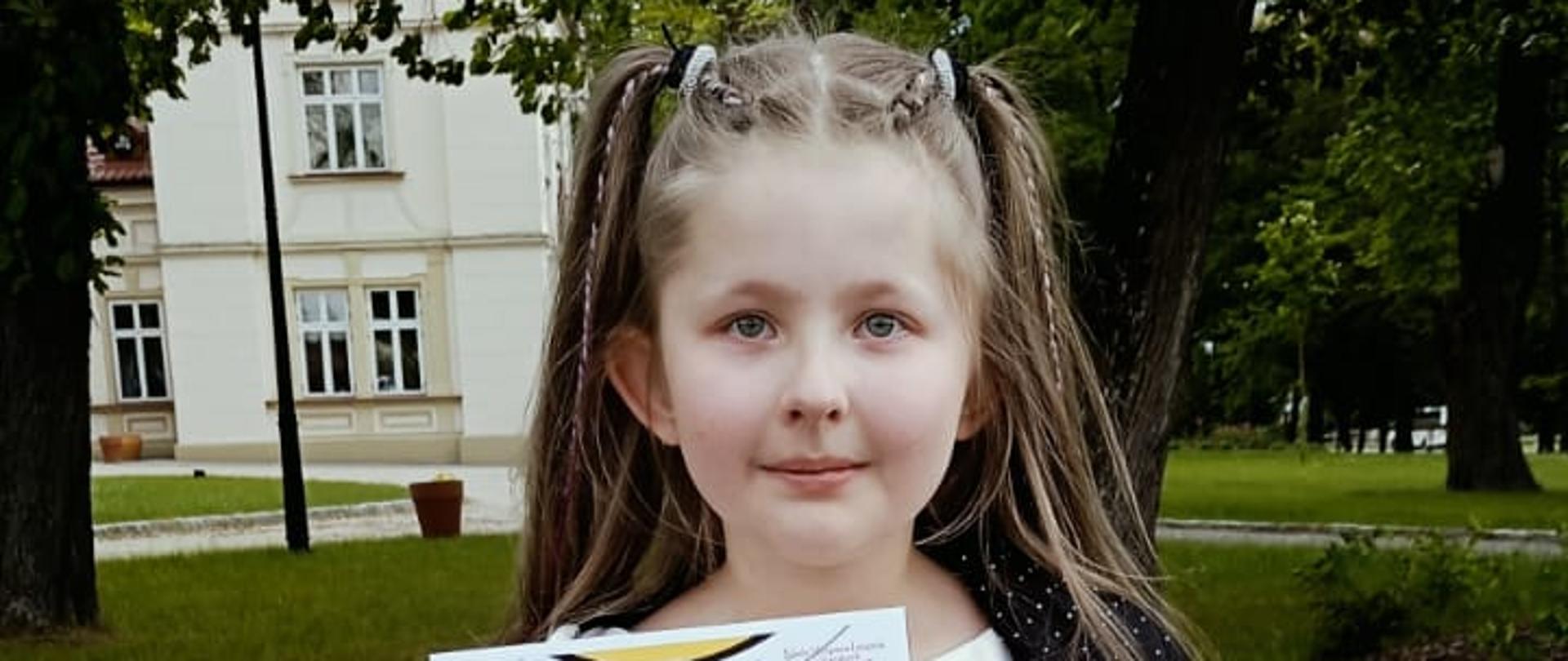 Mała dziewczynka stoi na trawie, trzymając w rękach dyplom z napisem „Wyróżnienie I stopnia” oraz papierową torebkę z nagrodą. Ubrana jest w białą sukienkę, czarną narzutkę w białe kropki i złote buty. Ma długie, jasne włosy upięte w dwa kucyki. W tle widać budynek w stylu klasycznym oraz drzewa. Dziewczynka z dumą prezentuje swój dyplom.