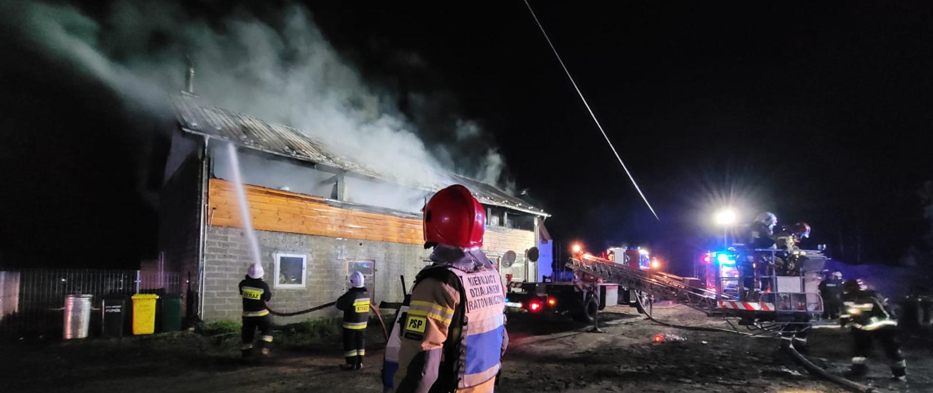 Strażak kierujący działaniami ratowniczymi obserwuje akcję. W tle widać palący się budynek gospodarczy oraz strażaków uczestniczących w akcji.