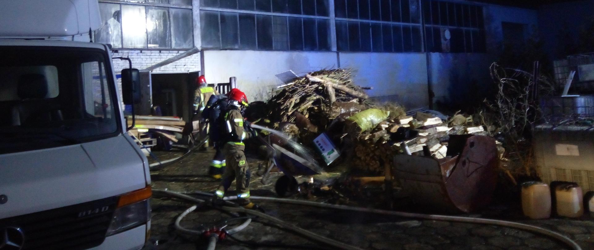 Zdjęcie przedstawia strażaka przelewającego wodą pogorzelisko. Z lewej strony widoczny jest biały samochód dostawczy. W tle magazyn w którym doszło do pożaru.