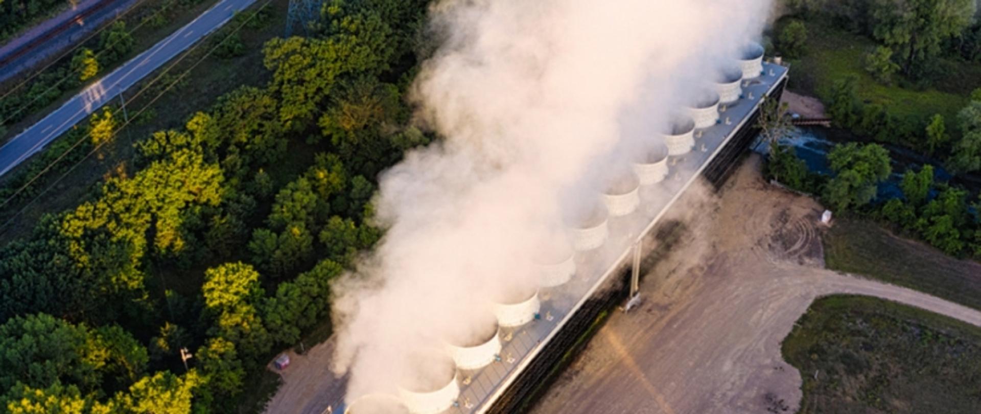 Zakład przemysłowy z wieżami chłodniczymi otoczony lasem; widok z góry z drogą asfaltową i ścieżką gruntową.