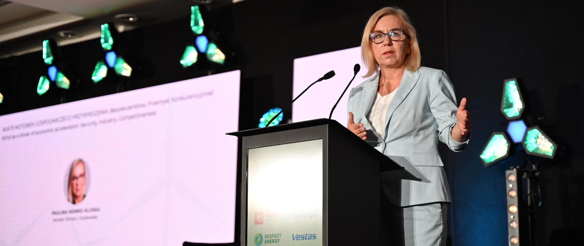
Ministra Paulina Hennig-Kloska - osoba w niebieskim garniturze - stoi za mównicą i przemawia. Po lewej stronie telebim ze zdjęciem twarzy minister oraz jej imieniem i nazwiskiem. 