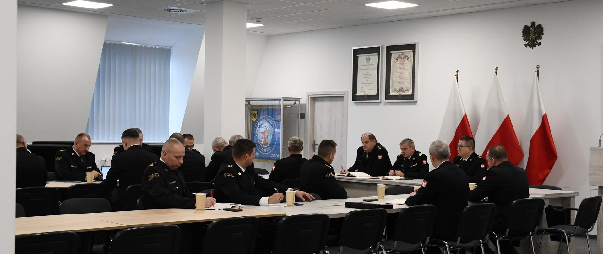 Strażacy siedzą za stołami za nimi znajdują się flagi Polski godło Polski sztandar Komendy Wojewódzkiej Państwowej Straży Pożarnej w Gdańsku oraz rota ślubowania strażaka.