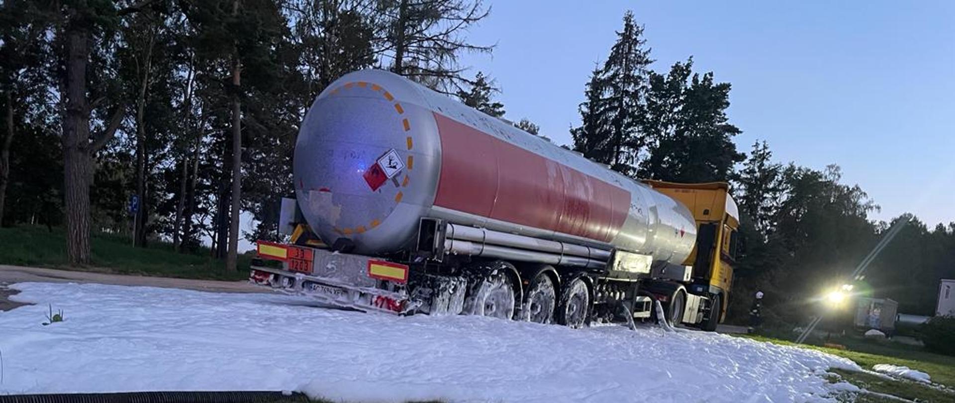 Zdjęcie przedstawia samochód ciężarowy z cysterną na parkingu w około cysterny rozlana jest piana ratownicza 