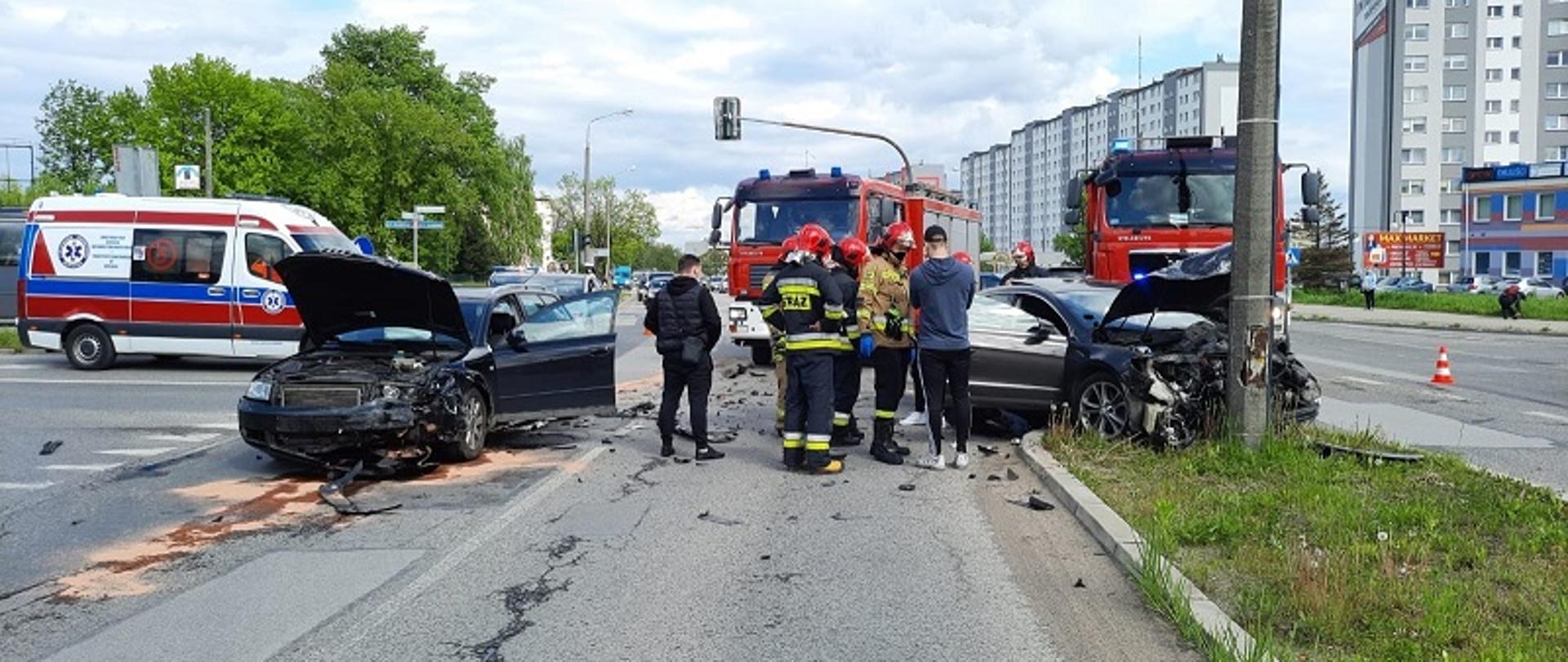 Na zdjęciu widzimy kolizję drogową dwóch samochodów osobowych: Audi A4 oraz Volkswagen Passat CC. Z lewej strony zdjęcia samochód Audi A4 ma uszkodzony prawy bok pojazdu. Po prawej stronie zdjęcia widzimy samochód Passat, który uderzył w przydrożny słup oświetleniowy. Pojazd ma uszkodzony przód. W dalszym tle zdjęcia widzimy Zespół Pogotowia Ratunkowego oraz strażackie pojazdy. Siła zderzenia dwóch pojazdów osobowych była tak duża, że na ulicy widać porozrzucane elementy karoserii obu pojazdów. Z samochodu Audi na pas drogowy wyciekły płyny eksploatacyjne, które zostały zneutralizowane przez strażaków sorbentem Compact.