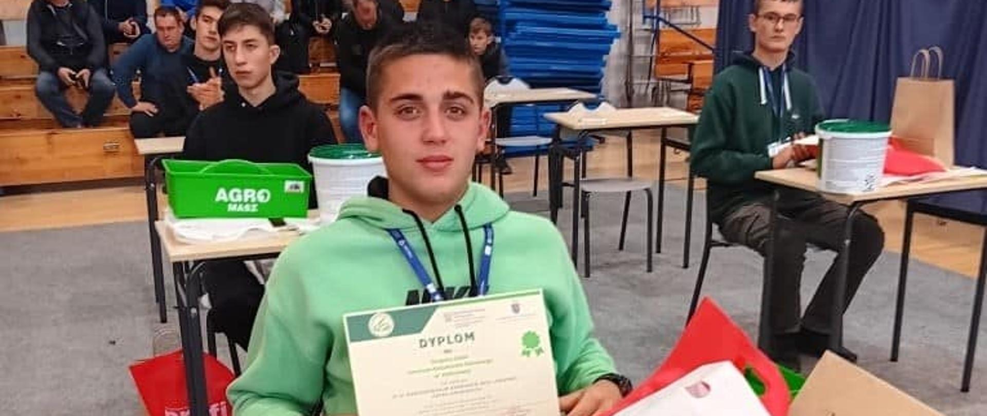 Młody mężczyzna w jasnozielonej bluzie siedzi przy stoliku i dumnie prezentuje otrzymany dyplom. W tle widać innych uczestników oraz trybuny sali gimnastycznej. Na stolikach znajdują się materiały promocyjne i puszki z logiem "Agro-Masz".