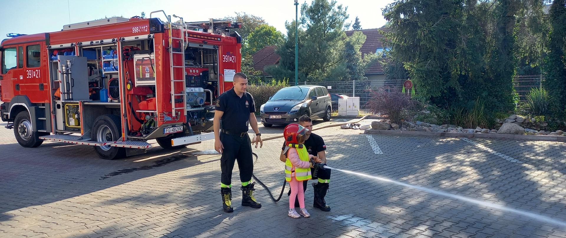 Widok dziecka ubranego w hełm strażacki, które w asyście dwóch strażaków podaje prąd wody. W tle samochód pożarniczy.