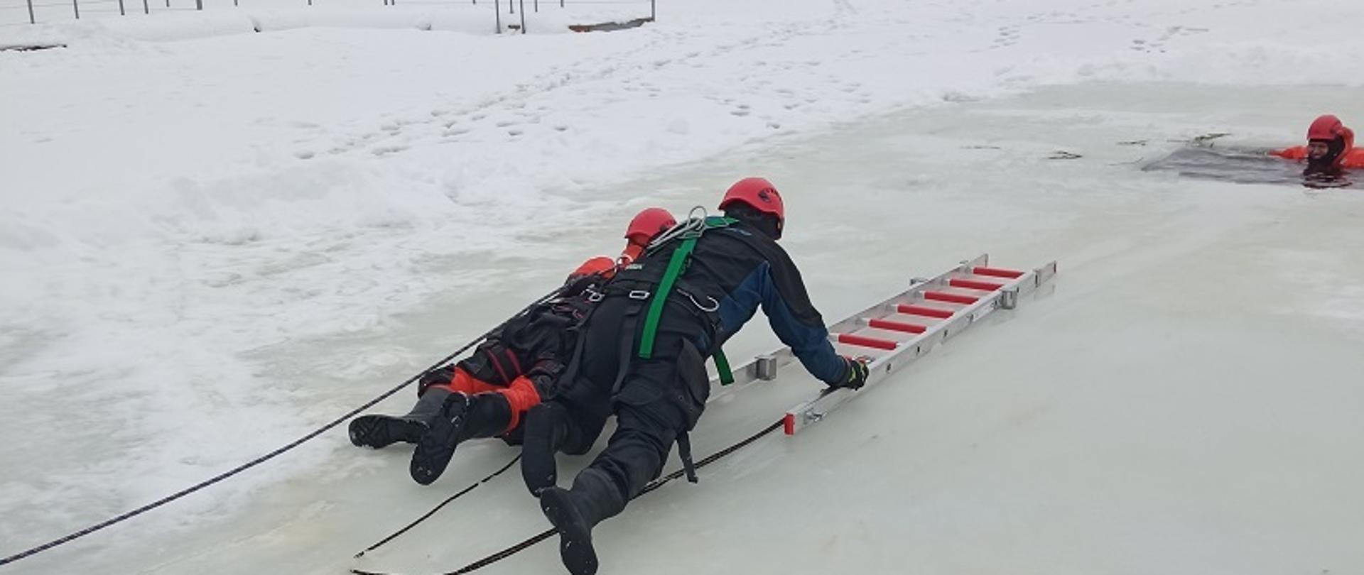 Zdjęcie obrazuje 2 ratowników przemieszczających się na czworakach na lodzie z drabiną w kierunku przerębla gdzie jest osoba w wodzie.