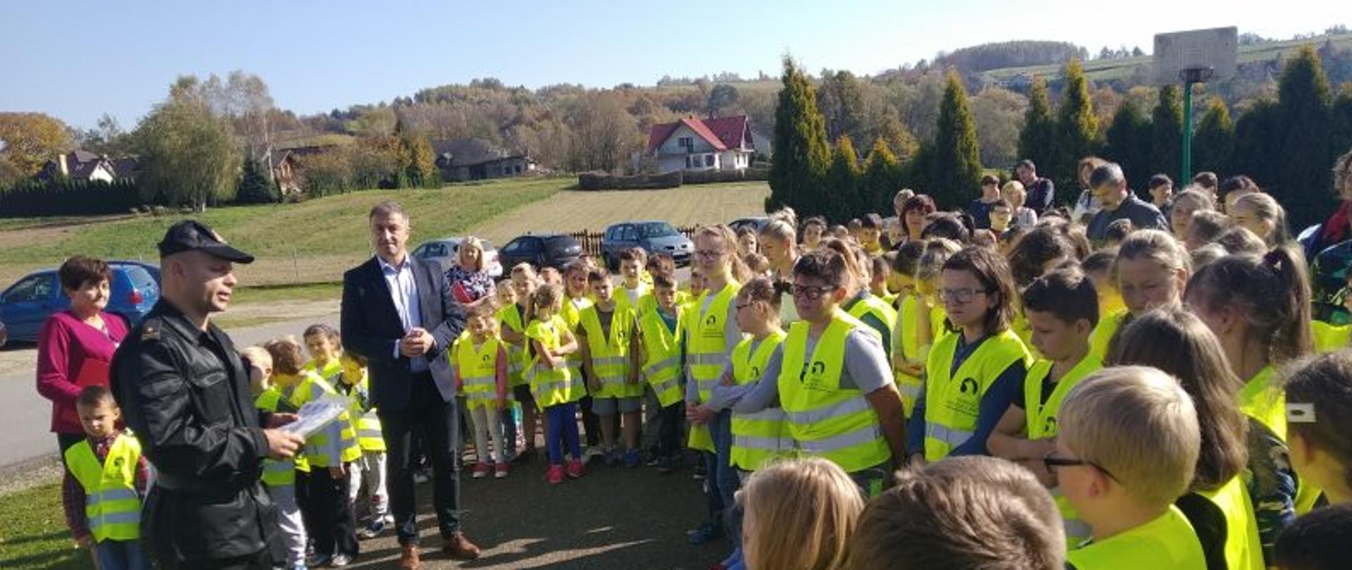 Na zdjęciu dzieci ubrane w żółte kamizelki z odblaskami wraz z nauczycielami i dyrektorem szkoły na środku komendant powiatowy Państwowej Straży Pożarnej w Jaśle wygłasza prelekcję w tle domy pagórki lasy