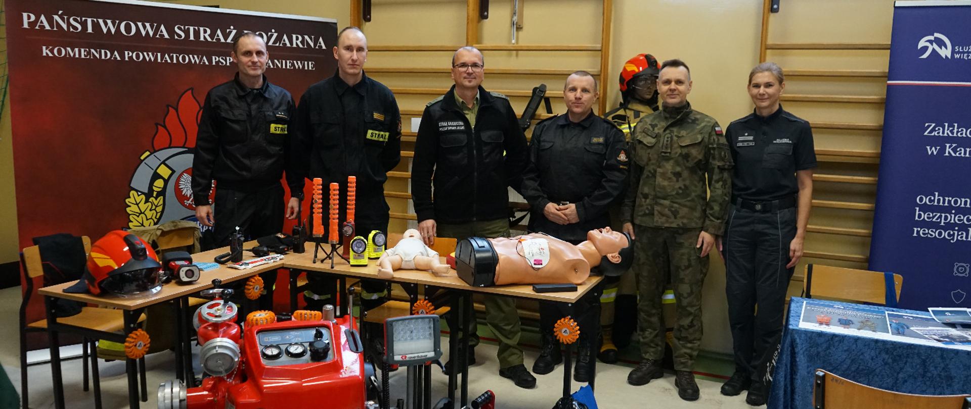 Zdjęcie zrobione na sali gimnastycznej. Przy drabinkach stoi grupa sześciu funkcjonariuszy. Z lewej strony baner Państwowej Straży Pożarnej. Przed nimi na podłodze oraz na dwóch stolikach sprzęt strażacki, czerwona pompa, torba medyczna, na stolikach latarki i manekiny.