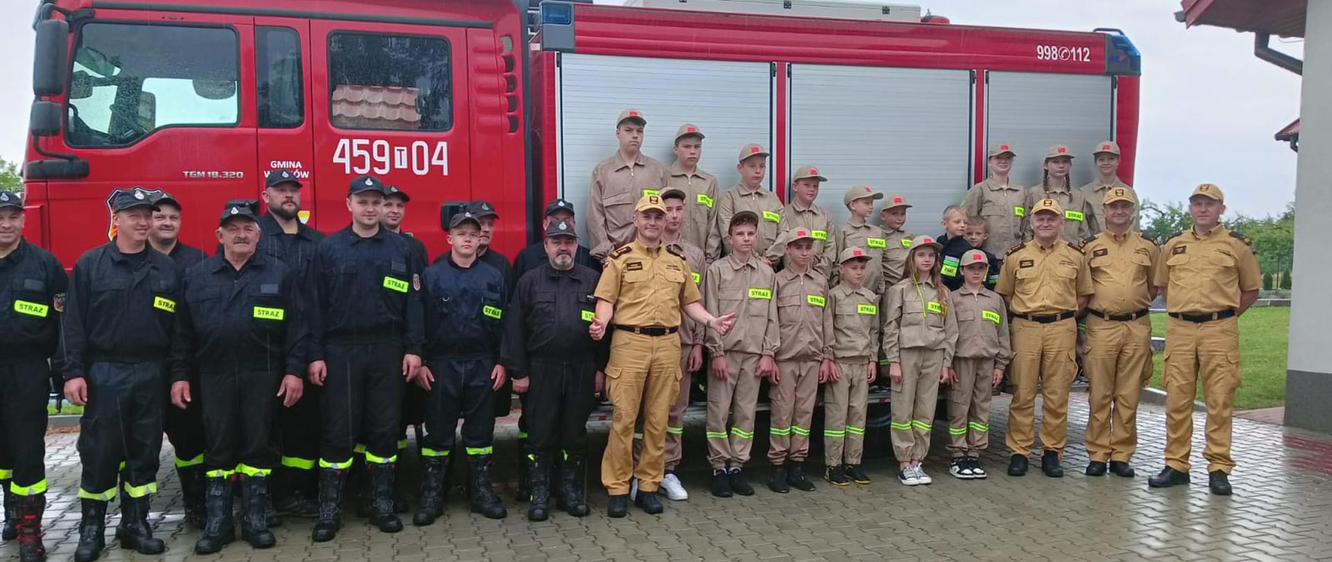 Na zdjęciu w aurze deszczowej widać strażaków OSP oraz dzieci z Młodzieżowych Drużyn Pożarniczych, którzy pozują do zdjęcia grupowego z Zastępcą Komendanta Wojewódzkiego PSP w Kielcach. W tle samochód gaśniczy z OSP Witosławice