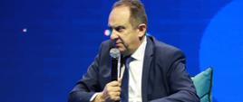 Wiceminister rozwoju i technologii Andrzej Gut-Mostowy na panelu poświęconemu turystyce podczas Europejskiego Kongresu Gospodarczego w Katowicach, wiceminister siedzi i trzyma w dłoni mikrofon