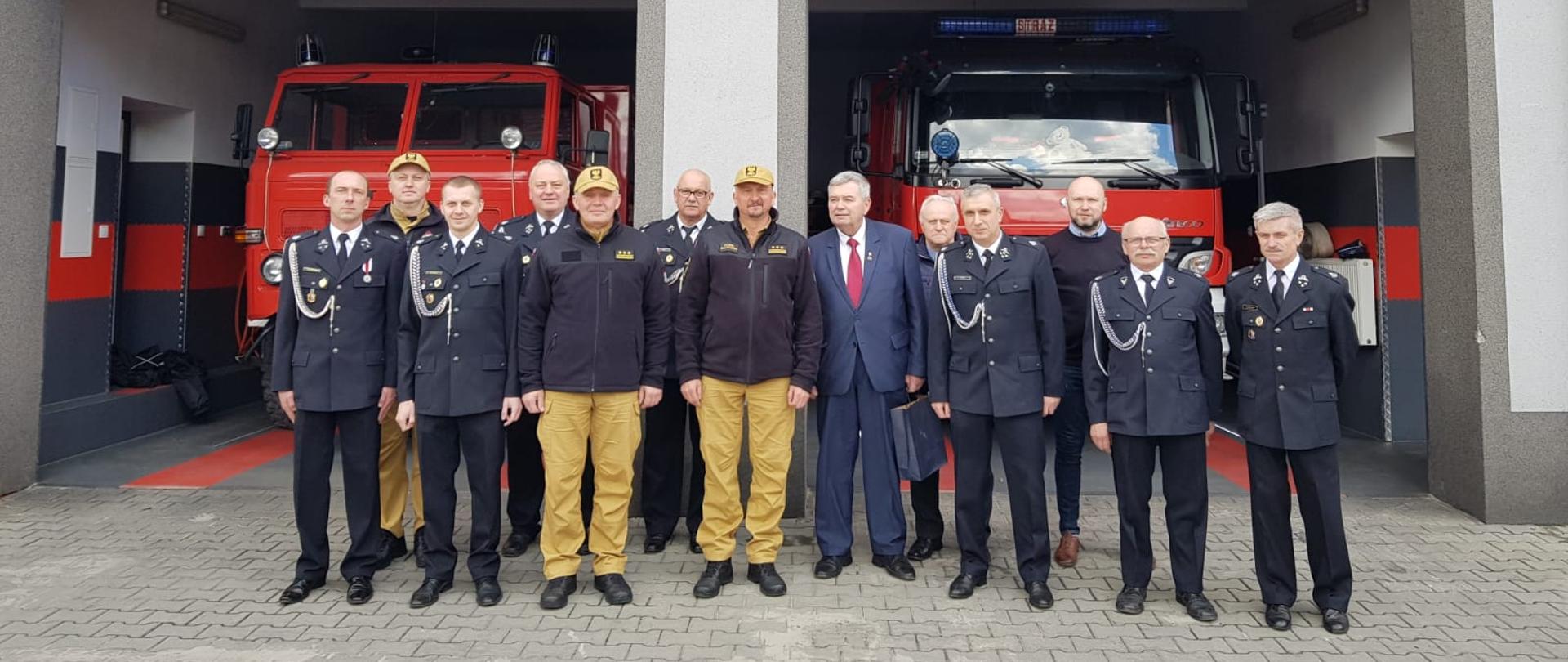 komendanci PSP, druhowie OSP oraz przedstawiciele samorządu pozują do zdjęcia przed jednostką OSP,, bramy garażowe są otwarte i widać 2 samochody pożarnicze