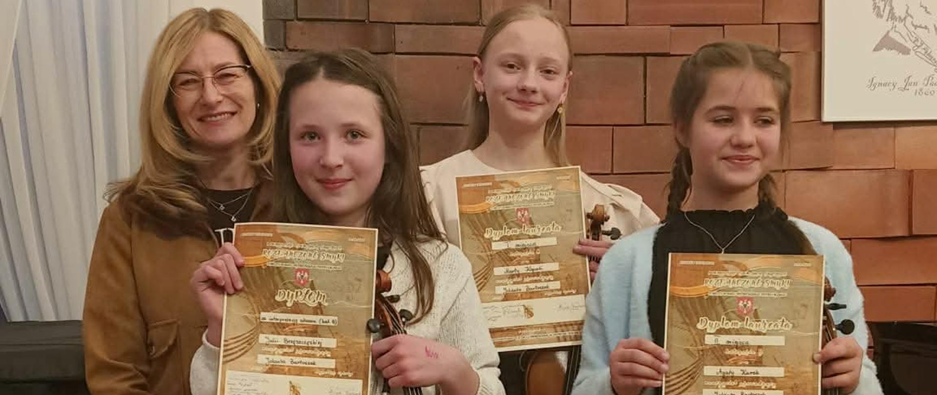 Na zdjęciu są trzy uczennice odświętnie ubrane. W ręku trzymają dyplomy i skrzypce. Obok nich stoi nauczycielka z klasy skrzypiec pani Jolanta Bartoszek.