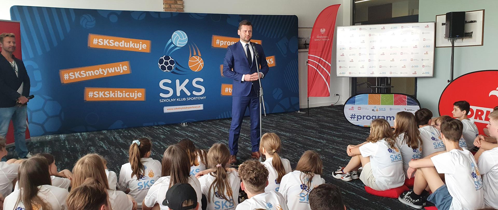 Minister Kamil Bortniczuk zachęcał uczniów do korzystania z oferty Programu Szkolny Klub Sportowy. Na zdjęciu minister Bortniczuk przemawia do siedzących na poduszkach uczniów w koszulkach SKS.