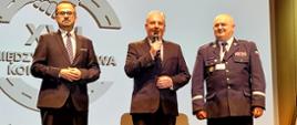 Trzech mężczyzn stoi na scenie, mężczyzna w środku trzyma mikrofon.