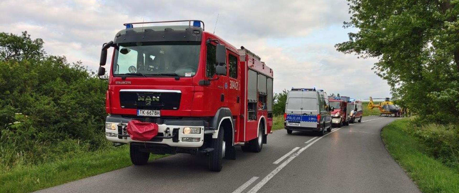 Na zdjęciu przedstawiono łuk drogi powiatowej, na którym stoją pojazdy służb w kolejności: straż pożarna, policyjny radiowóz, drugi samochód straży pożarnej, ambulans medyczny i śmigłowiec Lotniczego Pogotowia Ratowniczego. 