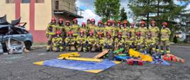 Zdjęcie przedstawia dużą grupę strażaków pozujących do fotografii na zewnątrz budynku, na placu szkoleniowym. W sumie jest około dwudziestu sześciu strażaków, zarówno mężczyzn, jak i kobiet. Wszyscy ubrani są w jednakowe mundury w kolorze piaskowym z czarnymi elementami i czerwone hełmy. Niektórzy strażacy stoją w tylnym rzędzie, a większość klęczy w dwóch rzędach z przodu. Wszyscy patrzą w stronę obiektywu.
Przed strażakami, na rozłożonej niebiesko-żółtej plandece, znajduje się różnorodny sprzęt ratowniczy. Można dostrzec między innymi rozpieracz hydrauliczny w czerwonej obudowie, węże strażackie, nosze, oraz inne narzędzia. Po lewej stronie zdjęcia widać rozbity samochód osobowy w jasnoszarym kolorze. Ma on zniszczony przód, powybijane szyby i otwarte drzwi. Fragmenty karoserii leżą obok pojazdu. W tle za strażakami znajduje się budynek o jasnej, beżowo-żółtej fasadzie z czerwonymi pasami. Budynek ma kilka pięter i charakterystyczną wieżę do ćwiczeń strażackich z balkonami i drabinkami. Po prawej stronie widać fragmenty innych budynków mieszkalnych oraz wysokie, zielone drzewa iglaste. Niebo nad nimi jest częściowo zachmurzone, z widocznymi jasnymi i ciemnymi chmurami.