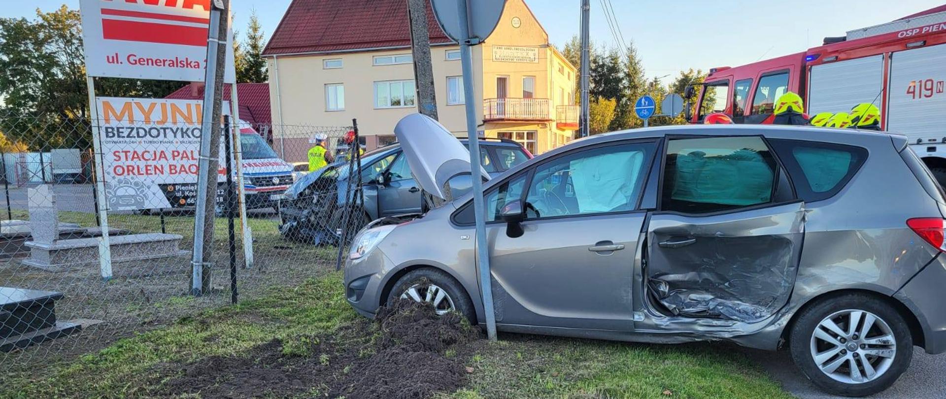 Na zdjęciu widoczna jest scena wypadku drogowego. Srebrny samochód osobowy uderzył bokiem w słup drogowy i wjechał na trawnik, uszkadzając ogrodzenie. Pojazd ma poważnie wgniecione drzwi od strony kierowcy oraz otwartą maskę. Na miejscu zdarzenia obecni są strażacy z oraz karetka pogotowia. Sytuacja wygląda na świeżo po kolizji, trwają działania służb ratunkowych.