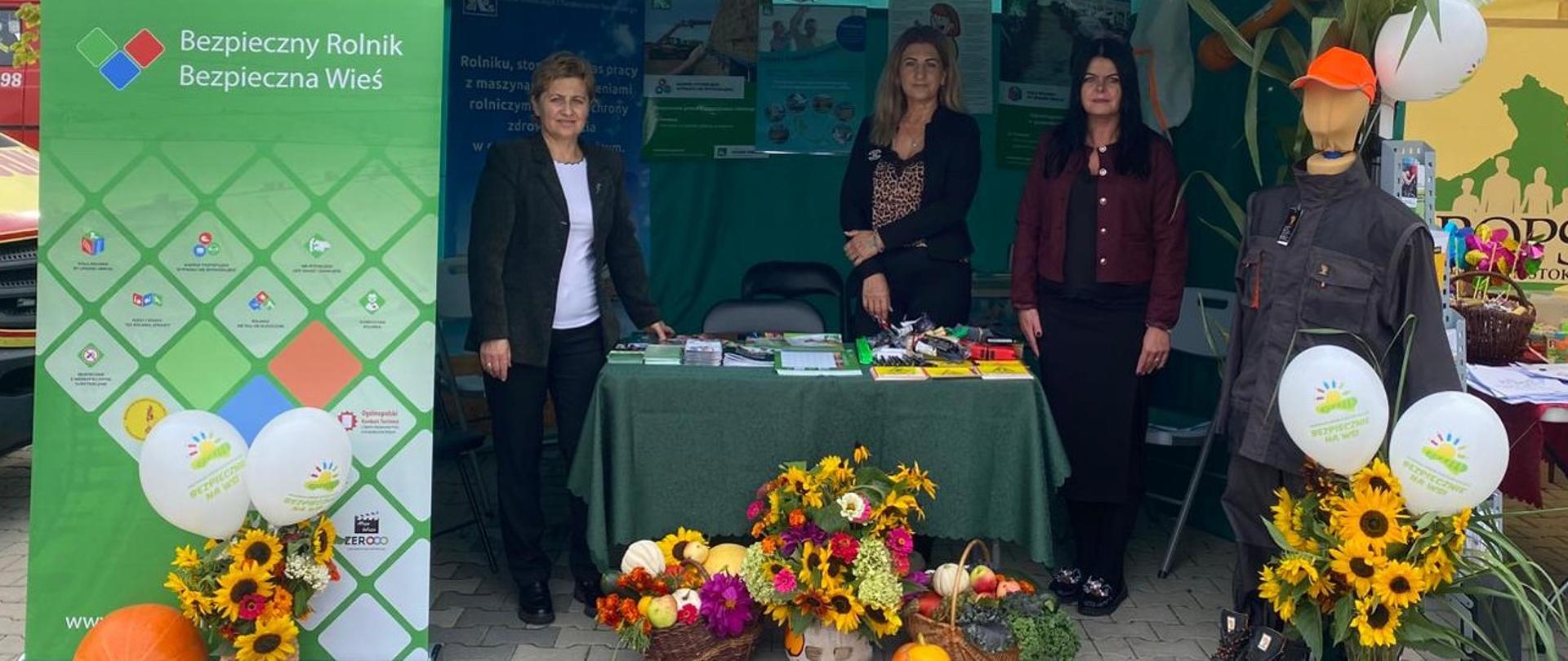 Trzy kobiety stoją pod namiotem informacyjnym KRUS przy stole z materiałami prewencyjnymi. Na pierwszym planie dekoracje dożynkowe — kosze z owocami, dynia i kwiaty. Po lewej widać roll-up z hasłem promocyjnym „Bezpieczny Rolnik, Bezpieczna Wieś”, po prawej manekin w odzieży roboczej i balony.