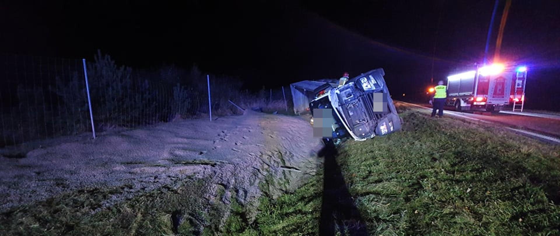 Kolizja na drodze krajowej. Auto ciężarowe leży w rowie na swoim prawym boku. Wokół rozsypany jest materiał przewożony pojazdem (kruszywo).Na jezdni stoi wóz straży pożarnej i policjant. Pora nocna. teren oświetlany jest a auta pożarniczego.