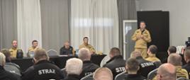 na sali widać siedzących tyłem strażaków a przed nimi przy stole prezydialnym siedzą funkcjonariusze PSP jeden stoi i pzremawia, w tle szare zasłony i biał firanki