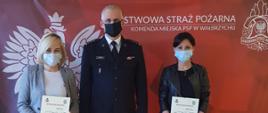 Zdjęcie przedstawia 3 osoby, z lewej kobieta w ciemnych spodniach i szarej marynarce, w środku galowo ubrany Komendant Miejski PSP w Wałbrzychu, z prawej strony kobieta w ciemnych spodniach i kurtce. Wszyscy stoją na tle czerwonego baneru