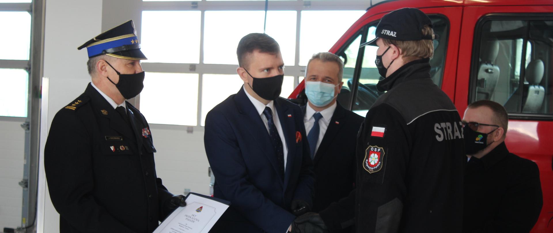 Na zdjęciu Minister Łukasz Schreiber oraz kujawsko-pomorski komendant wojewódzki przekazują pojazd strażakom z OSP Bydgoszcz-Fordon. Wręczają akt przekazania pojazdu oraz kluczyki Komendantowi tej straży. W tle goście m.in. wojewoda kujawsko-pomorski, przedstawiciele starostwa. Za nimi pojazd pożarniczy. Wydarzenie ma miejsce w garażu nowej remizy w Świeciu.