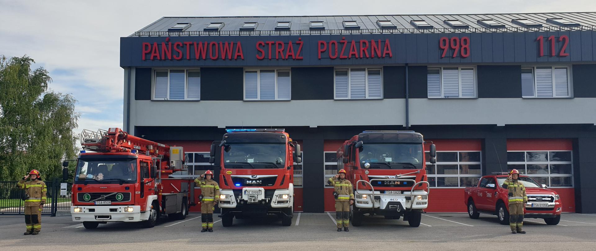 Zdjęcie przedstawia strażaków w ubraniach bojowych stojących na baczność w szeregu oddających hołd żołnierzom Powstania. W tle widać budynek Jednostki Ratowniczo- Gaśniczej a także samochody pożarnicze z włączonymi światłami drogowymi i świetlnymi.