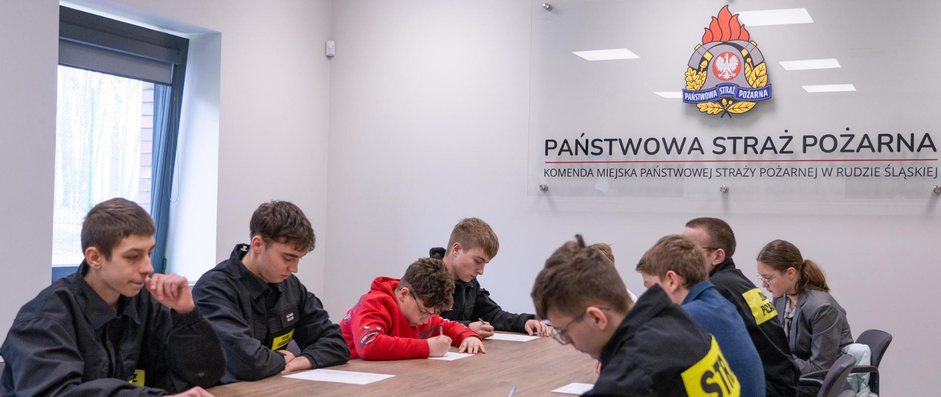 Grupa uczniów szkół podstawowych i ponadpodstawowych siedzi przy stole w sali Komendy Miejskiej PSP w Rudzie Śląskiej i wypełnia test w ramach OTWP; na ścianie widoczne jest logo Państwowej Straży Pożarnej.