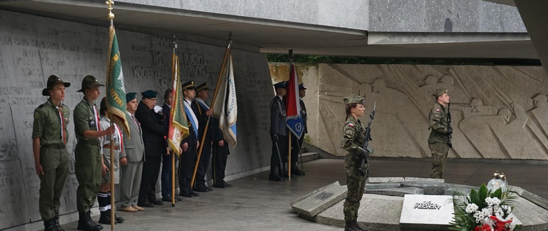 poczty sztandarowe stoją pod betonowym pomnikiem, przed nimi warta dwóch osób w mundurach wojskowych i z karabinami, przed wartą leży biało czerwony wieniec oraz palą się znicze