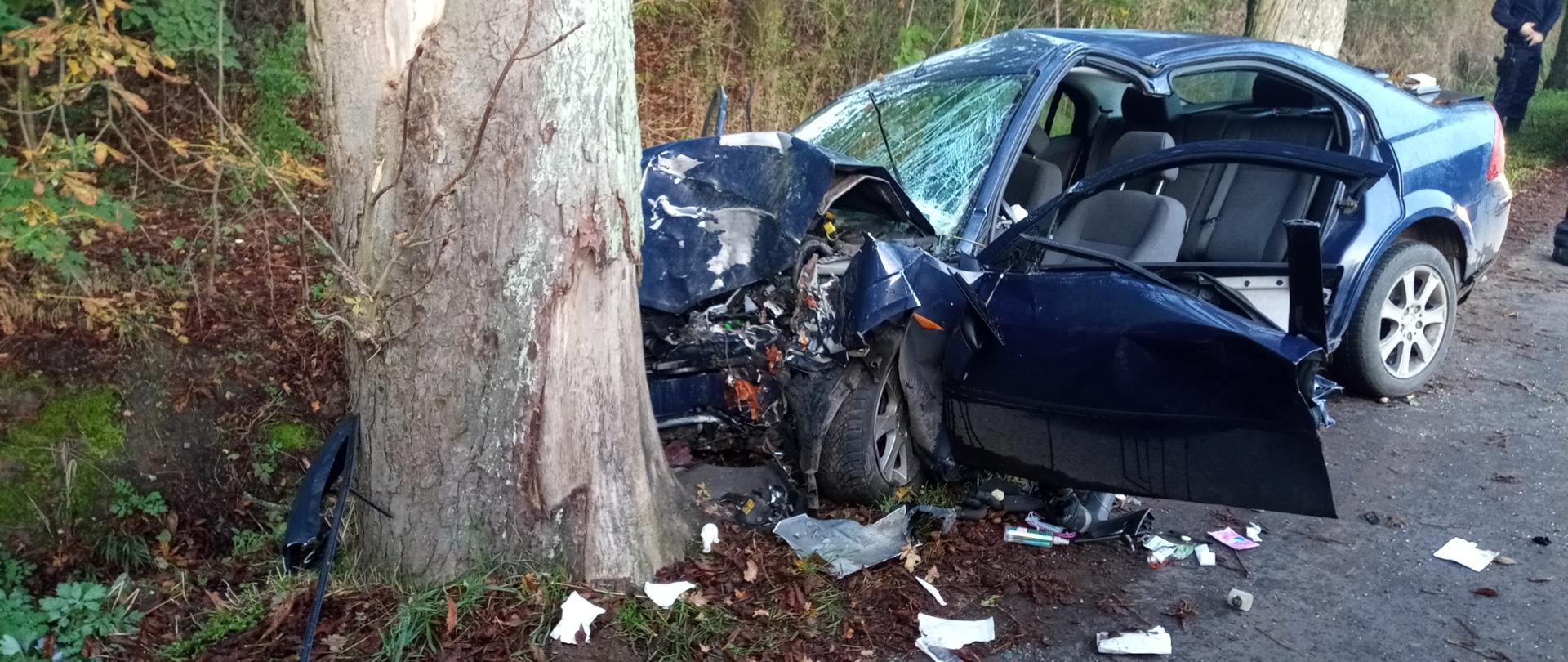 Auto osobowe stoi przy drzewie z widocznymi cięciami wykonanymi przez strażaków oraz ma zniszczony przód po uderzeniu w drzewo