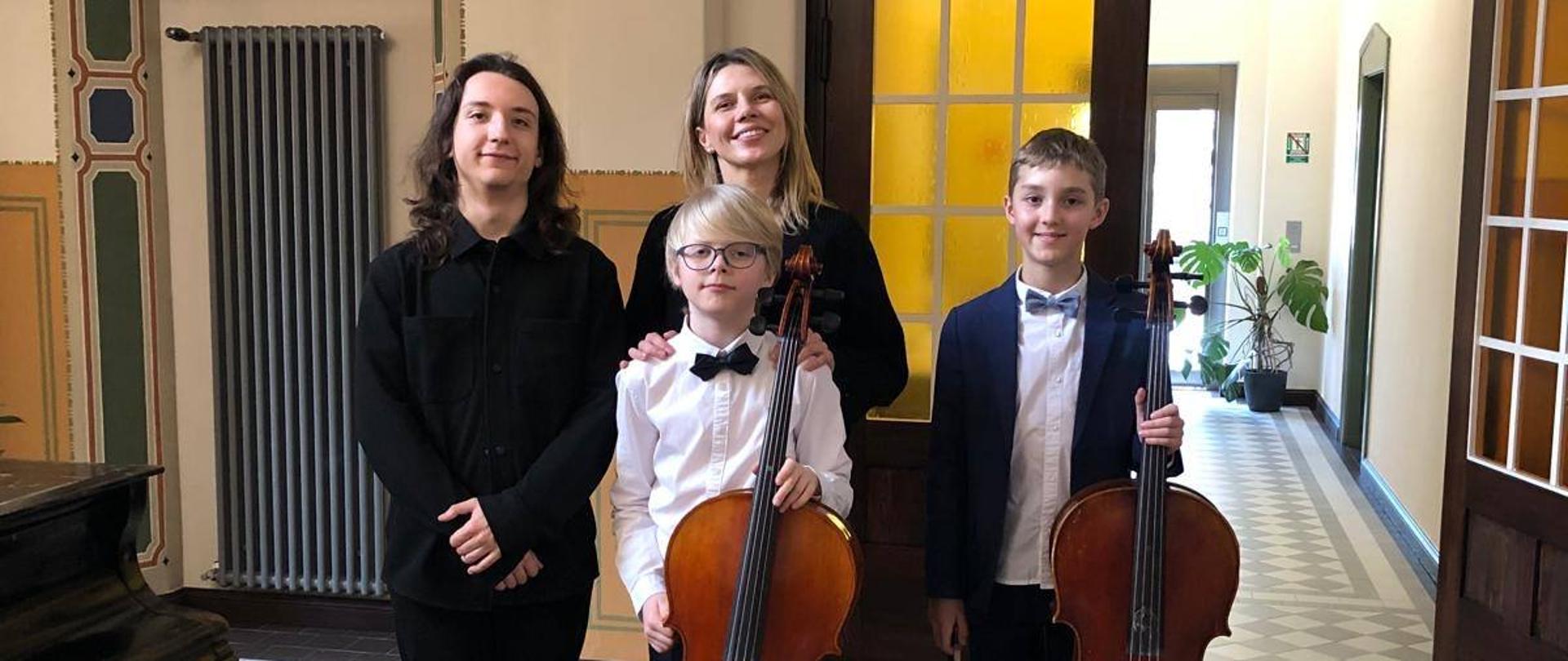 Maria Staśkowiak i Ryszard Chwalisz wraz dwójką uczniów pozują na stojąco. Dzieci trzymają wiolonczele.