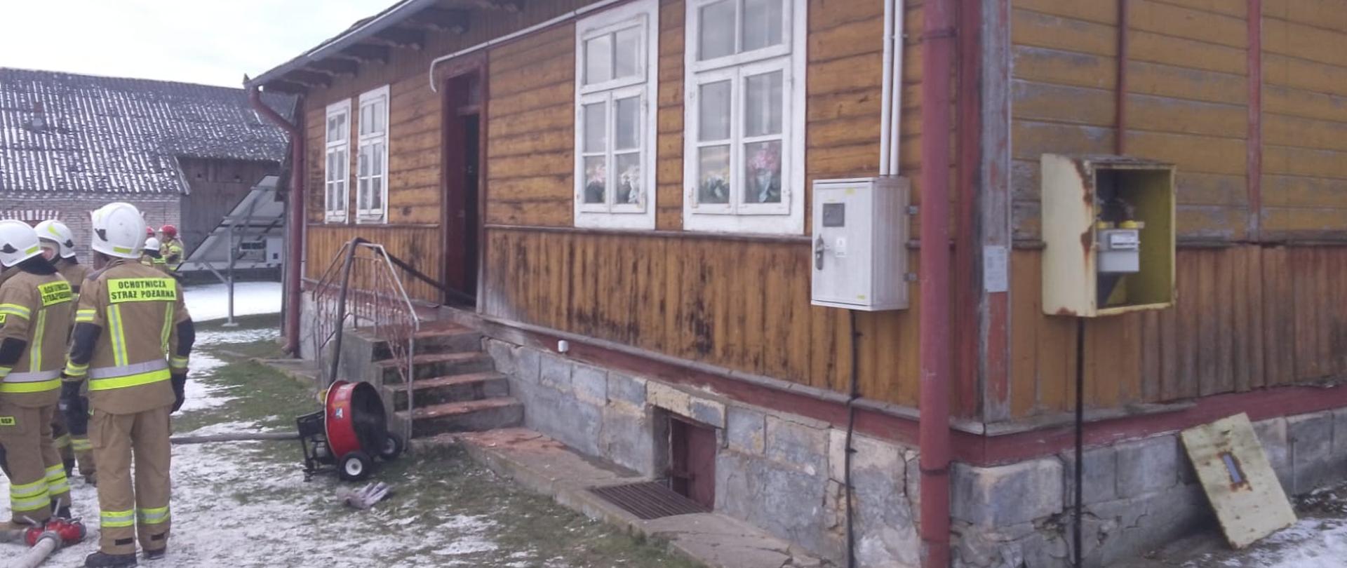 Po prawej stronie widać drewniany dom z oknami i drzwiami, po lewej stronie koło schodów stoi wentylator oddymiający, po lewej stronie stoją strażacy w piaskowych ubraniach i hełmach, obok nich przebiega linia gaśnicza 