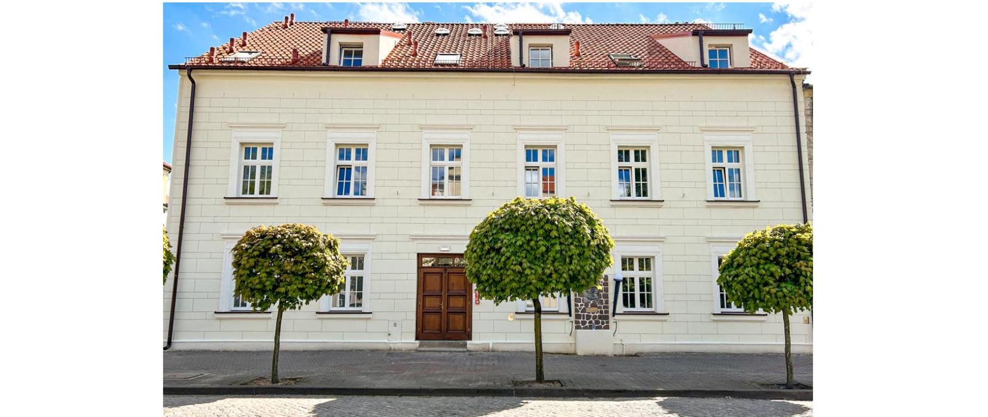 Na zdjęciu widoczny jest trzykondygnacyjny budynek oraz trzy drzewa rosnące przy brukowanej drodze. Na parterze budynku znajduje się pięć okien oraz drewniane drzwi wejściowe. Na prawo od nich widoczny jest pomnik umiejscowiony na ścianie budynku. Na pierwszym piętrze jest 6 okien, a na drugim 3 okna wykuszowe oraz trzy dachowe. Dach budynku pokryty jest dachówką w kolorze ceglanym. Natomiast elewacja ma śmietankowo biały kolor.
