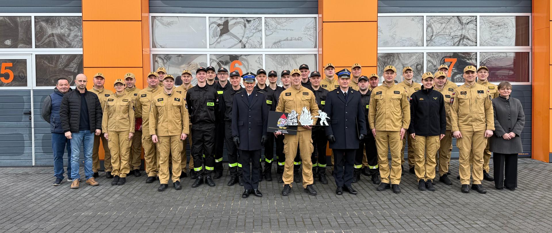 Zdjęcie grupowe wykonane przed bramami garażowymi komendy. W centrum stoi żegnany funkcjonariusz w otoczeniu kadry kierowniczej oraz strażaków. Trzyma pamiątkowy upominek. Wszyscy ustawieni są w kilku rzędach do wspólnej fotografii.