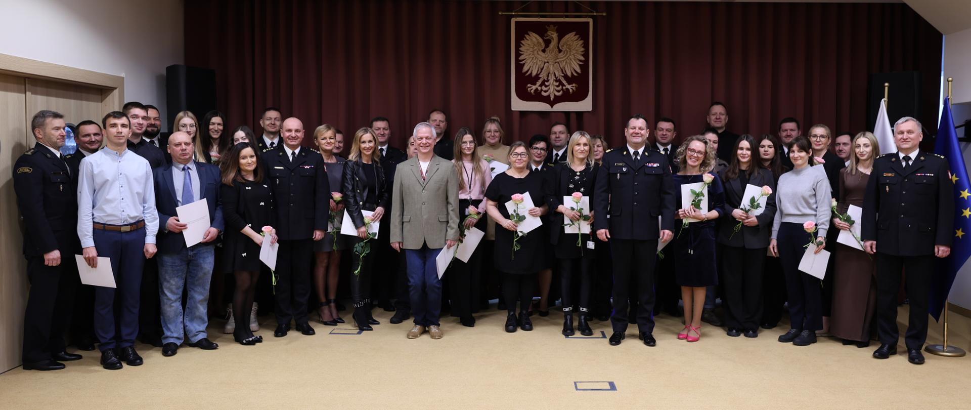 Zdjęcie grupowe wszystkich uczestników uroczystości zrobione na sali konferencyjnej. Sala jest kremowa, z beżową wykładziną i bordowymi kotarami.