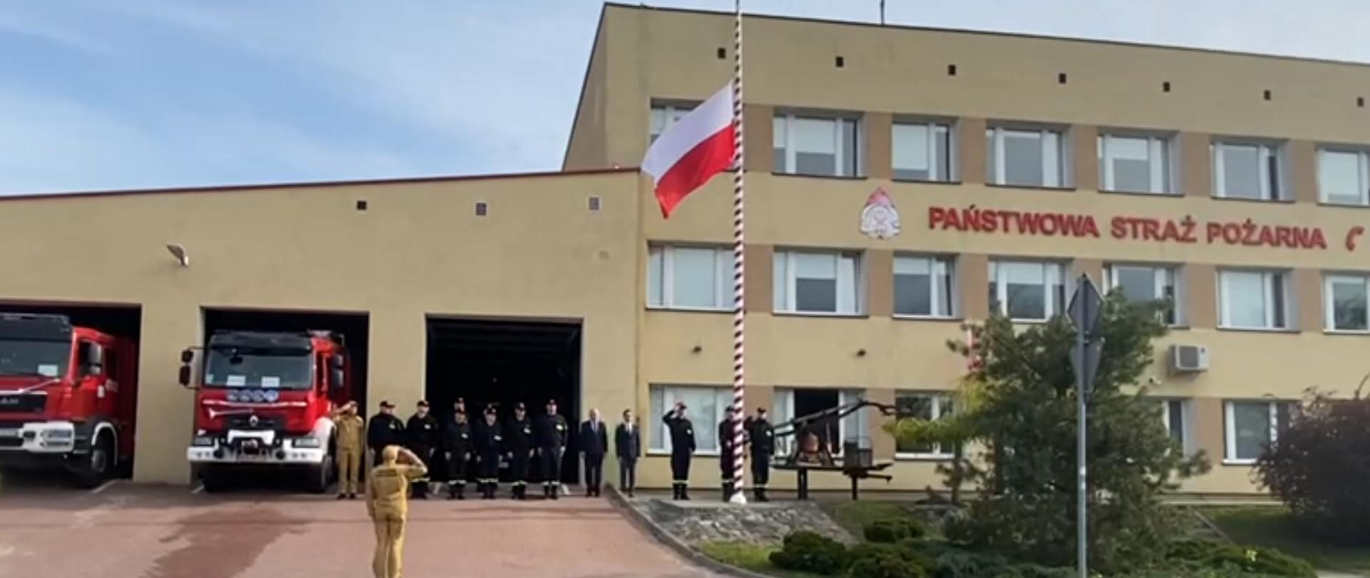 Zdjęcie zrobione przed budynkiem komendy oraz garaży. Budynek koloru żółtego. Przed budynkiem biało-czerowny maszt z zawieszoną flagą wciągniętą na ¾ wysokości. Strażacy ustawieni w szeregu.